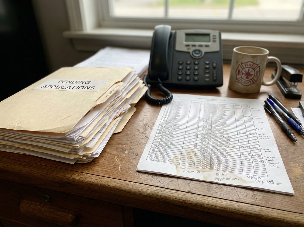 Cluttered desk with a pending applications folder, spreadsheets and a phone